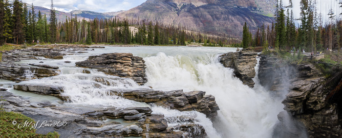 Panoramic view of a waterfall cascading over rocky cliffs, surrounded by lush green trees and majestic mountains in the background.