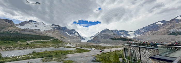 A panoramic view of the Icefields Parkway in the Canadian Rockies, showcasing majestic mountains, a glacier, and a cloudy sky.