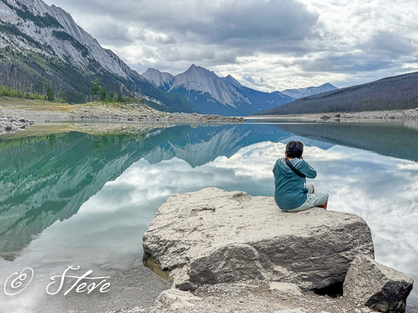 A person sitting on a rock by a serene lake, surrounded by mountains, with a cloudy sky reflecting on the water's surface.
