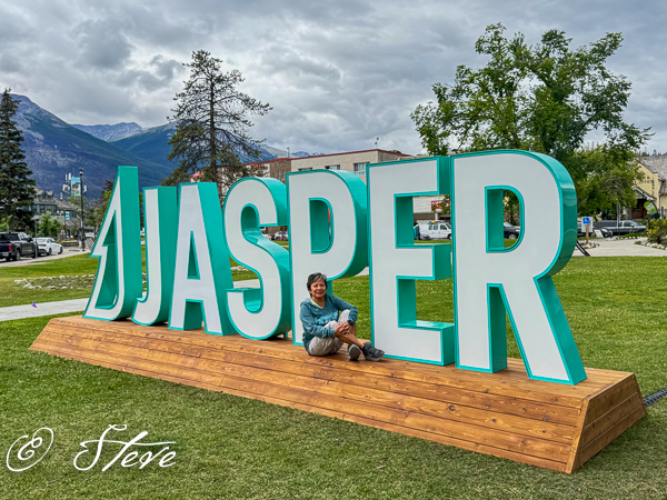 A person sitting on a wooden platform next to a large colorful sign that reads 'JASPER', with lush greenery and mountains in the background.