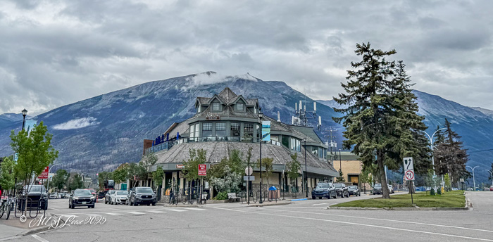 A view of a town square with buildings and shops, framed by mountains in the background under a cloudy sky.