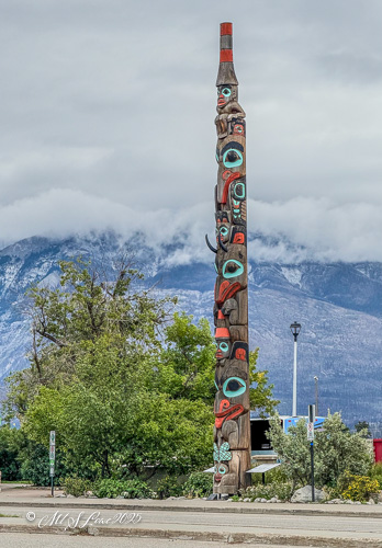 A tall totem pole with intricate carvings and vibrant colors, standing in an open area with mountains and cloudy skies in the background.