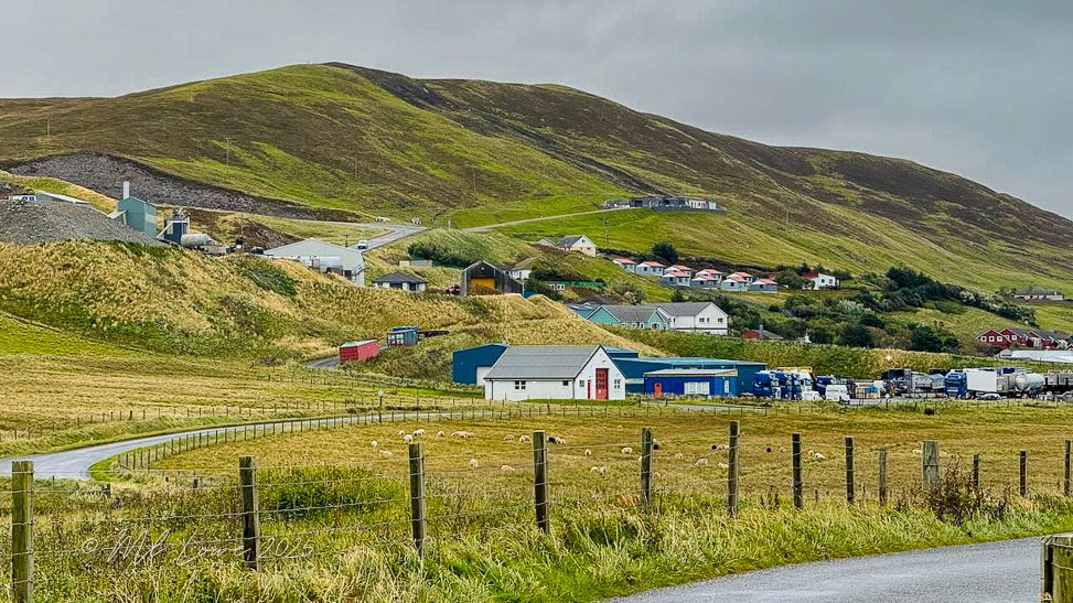 A scenic view of a rural landscape featuring rolling hills, a winding road, and small buildings with a backdrop of cloudy skies.