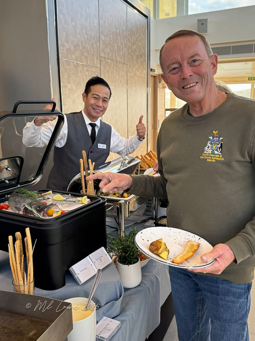 A man smiling while holding a plate with food, standing next to a server giving a thumbs up, in a dining setting with a buffet display.