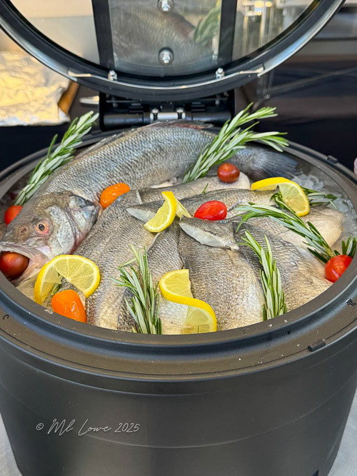 A display of fresh fish arranged with lemon slices, cherry tomatoes, and rosemary in a black container.