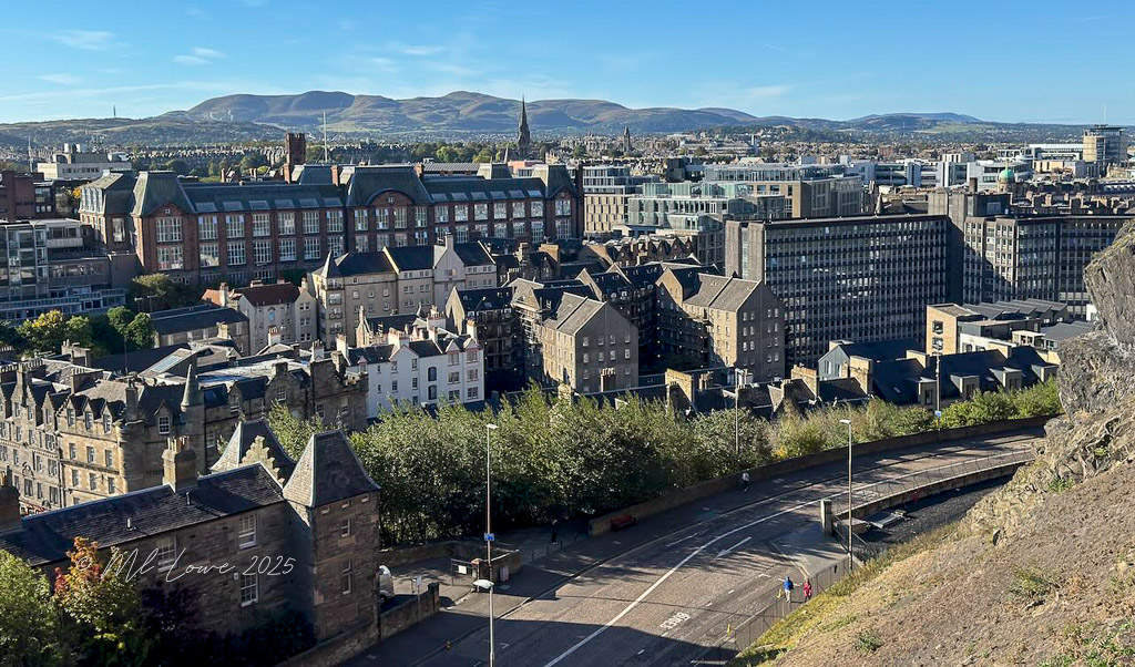 A panoramic view of Edinburgh, Scotland, showcasing a mix of historical buildings and modern architecture, with hills in the background under a clear blue sky.