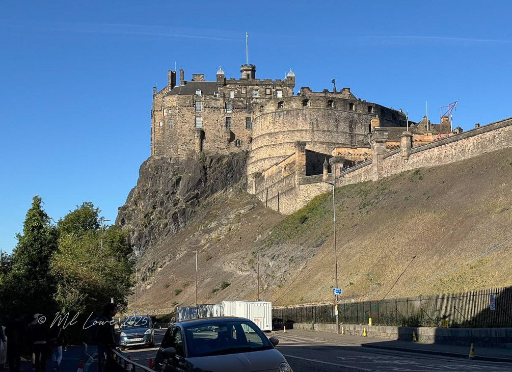A view of Edinburgh Castle perched on a rocky hill, with green trees and a clear blue sky in the background.