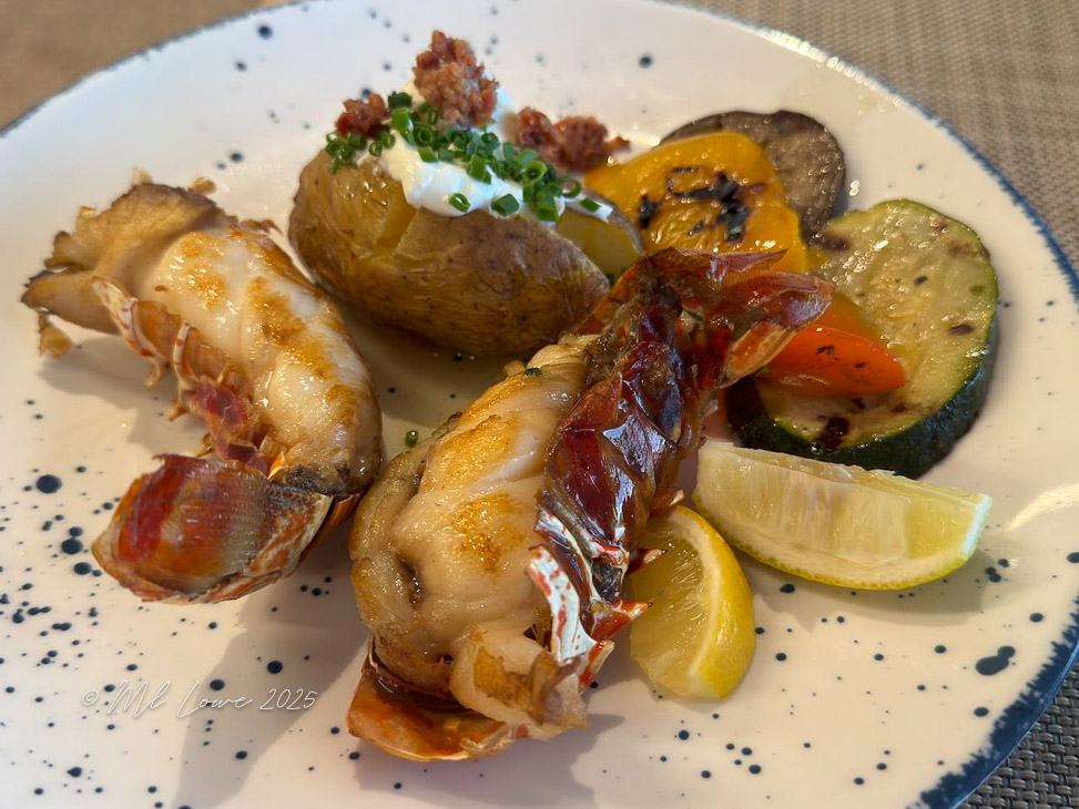 A plate of freshly cooked seafood featuring grilled lobster tail, a baked potato topped with sour cream and chives, and a medley of colorful roasted vegetables, garnished with lemon wedges.