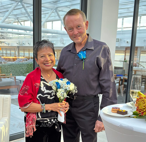 A woman holding a bouquet of flowers with a man beside her, both smiling in a well-lit indoor setting.