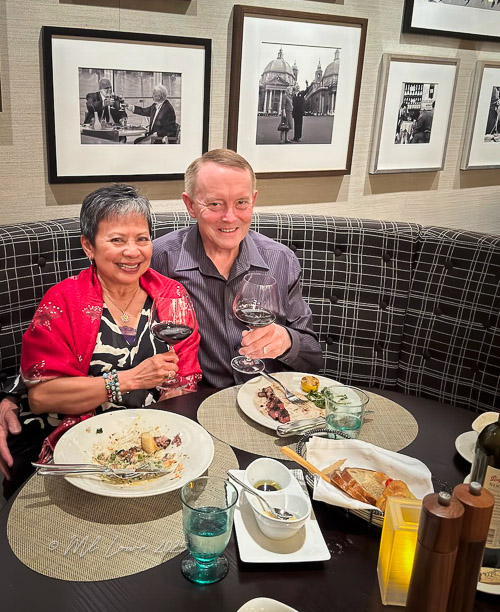 A couple toasting with glasses of wine at a restaurant table, surrounded by empty plates and framed photographs on the wall.