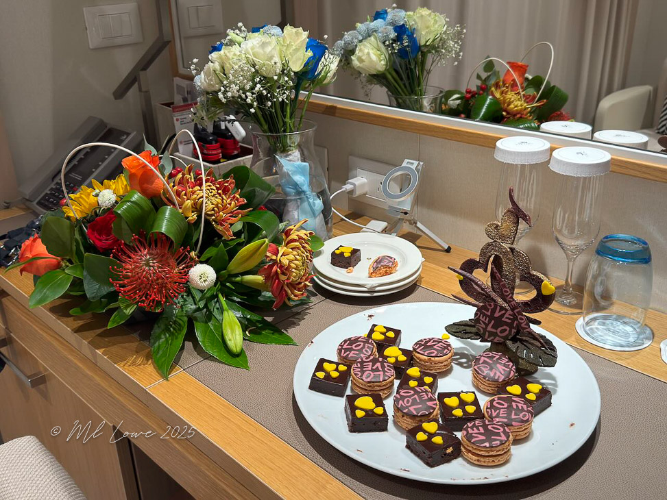 An arrangement of colorful flowers with a variety of blooms placed on a table, alongside a platter of decorated desserts including chocolate treats and cookies with love-themed designs.