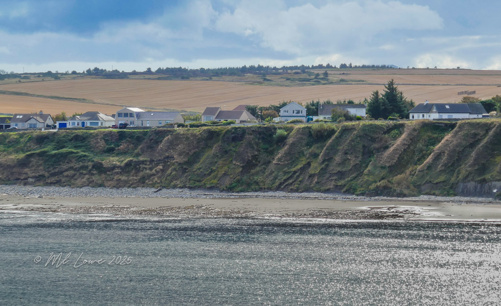 A coastal view featuring houses perched on a cliff with agricultural fields in the background under a cloudy sky.