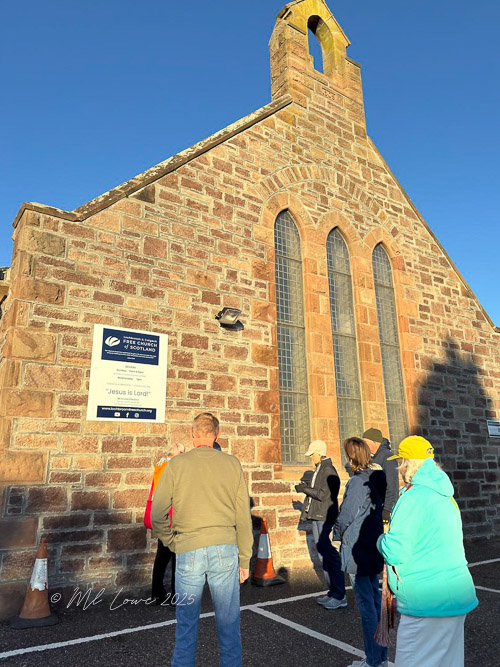 A group of people standing in front of a stone church, observing a sign on the building's exterior. The church features tall arched windows and a bell tower.