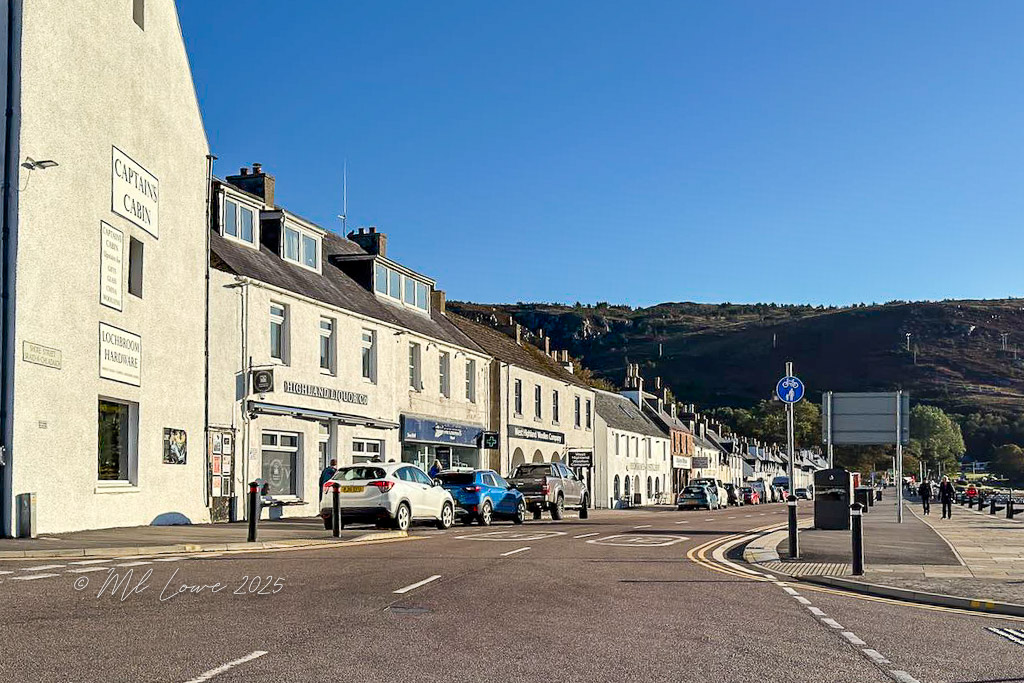 A street view of Ullapool, Scotland, featuring white buildings including a shop named 'Captain's Cabin' and parked cars along the road under a clear blue sky.
