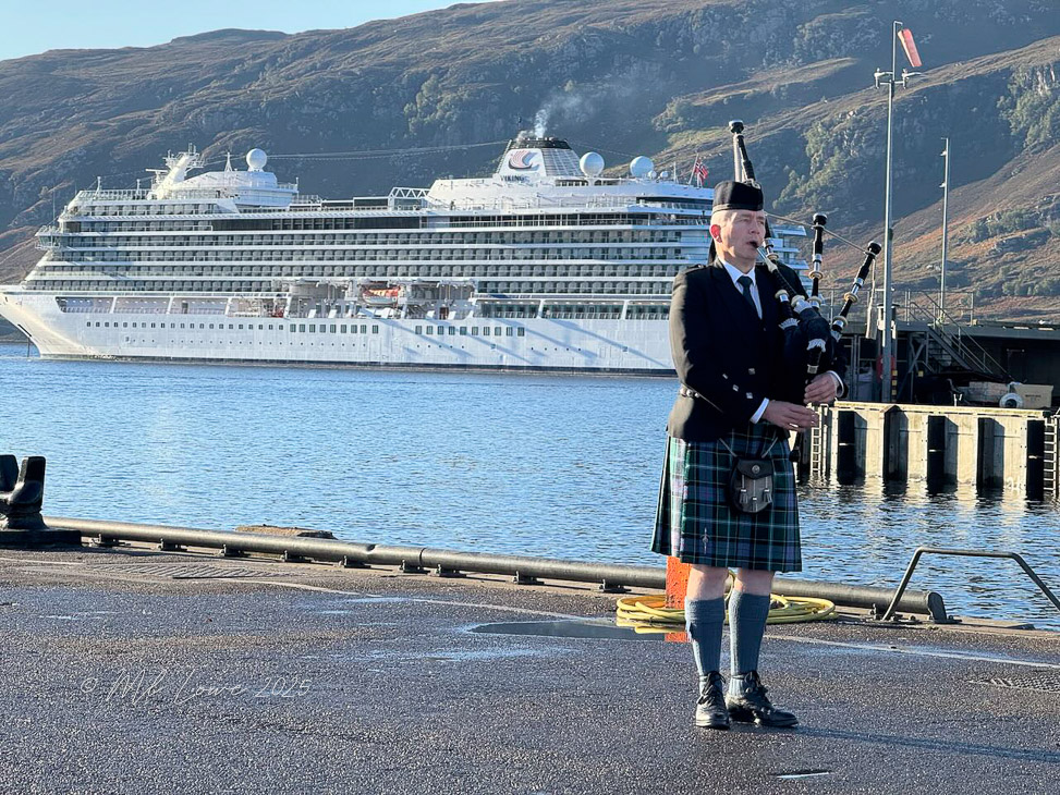 A man in a kilt playing bagpipes near a harbor with a large cruise ship in the background, surrounded by hills.