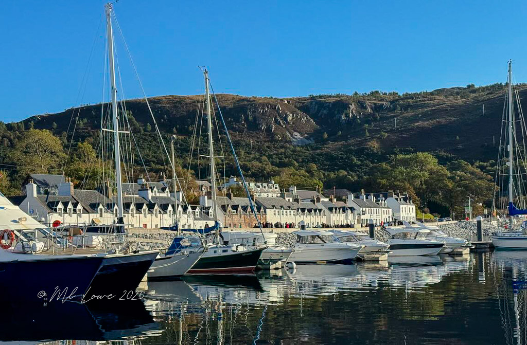 A picturesque harbor scene featuring several boats docked with a backdrop of quaint buildings and rolling hills under a clear blue sky.