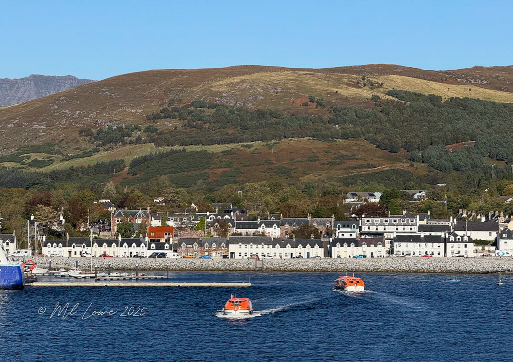 A scenic view of Ullapool, a fishing town in the Scottish Highlands, featuring colorful buildings along the waterfront and hills in the background, with boats navigating the water.