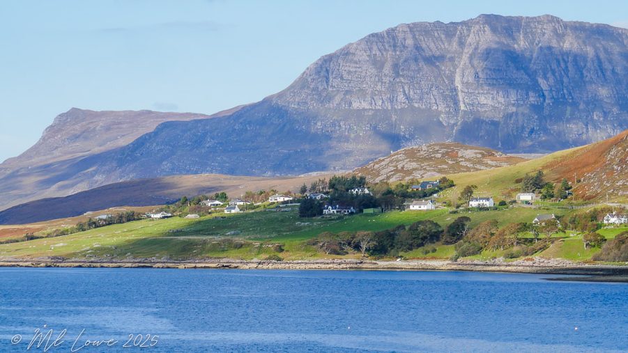 A scenic view of a coastal village at the base of a mountain, featuring green hills and blue water under a partly cloudy sky.