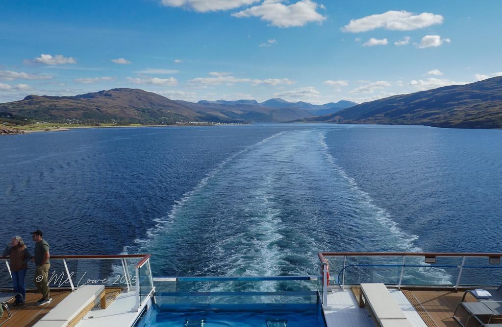 View from a cruise ship showing a tranquil sea and mountainous landscape under a blue sky.