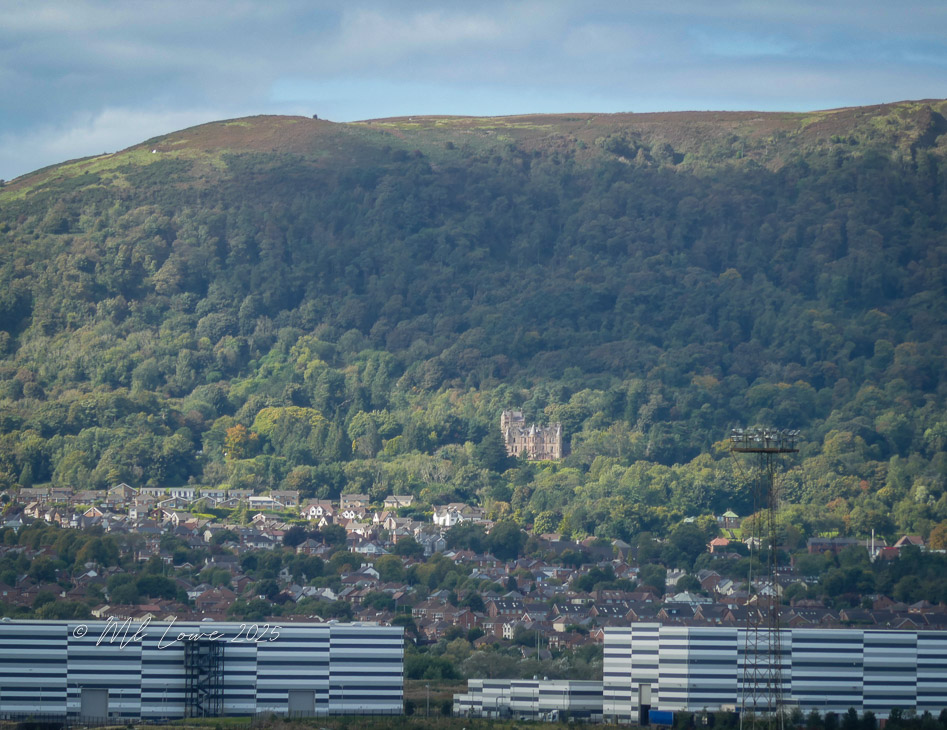Distant view of a castle nestled in a green forested hillside, with houses in the foreground and modern structures at the bottom of the image.