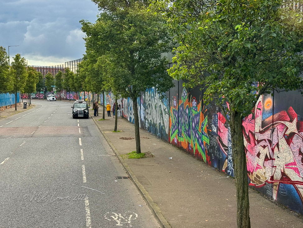 A street view in Belfast, Northern Ireland, featuring a wall covered in colorful political murals, with trees lining the road and a taxi parked nearby.
