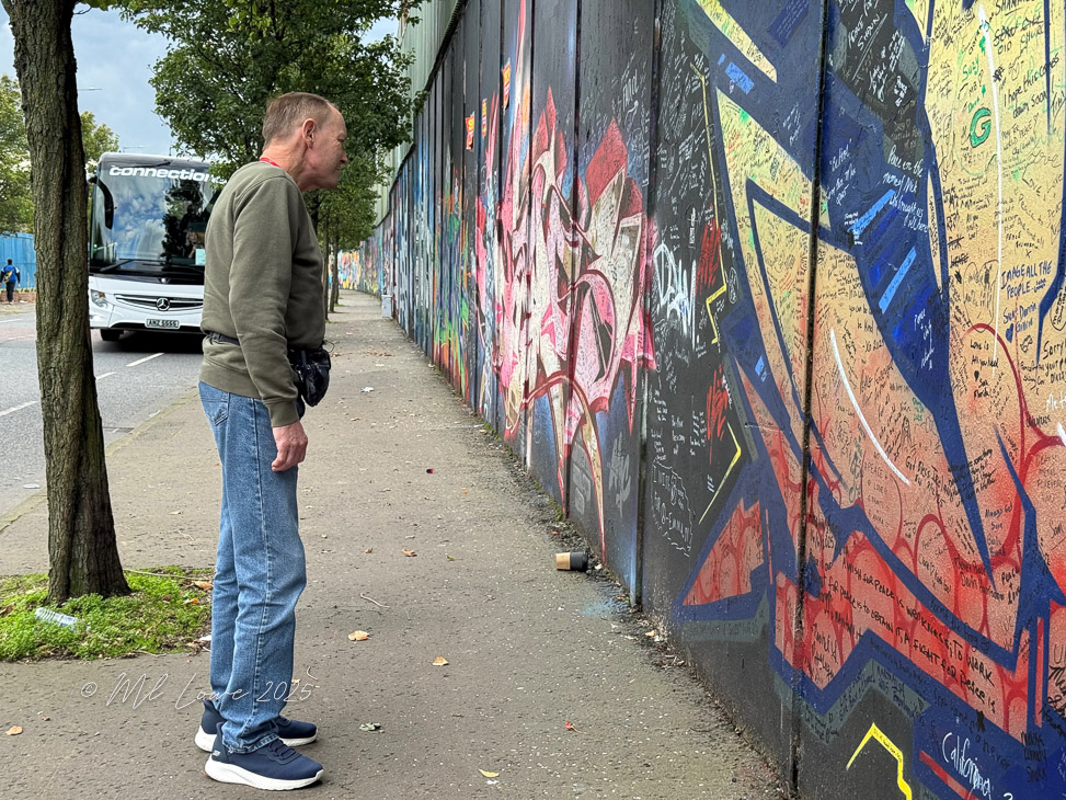 A person standing in front of a colorful graffiti wall filled with messages and drawings, with a tour bus visible in the background.