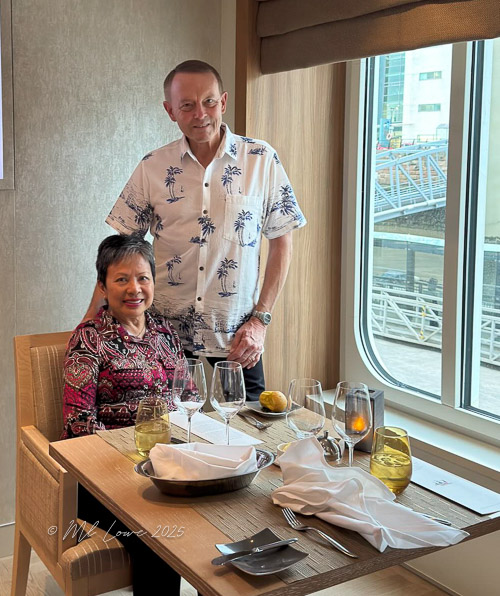 A man and a woman sitting at a dining table with a window view, enjoying a meal with elegant table settings.