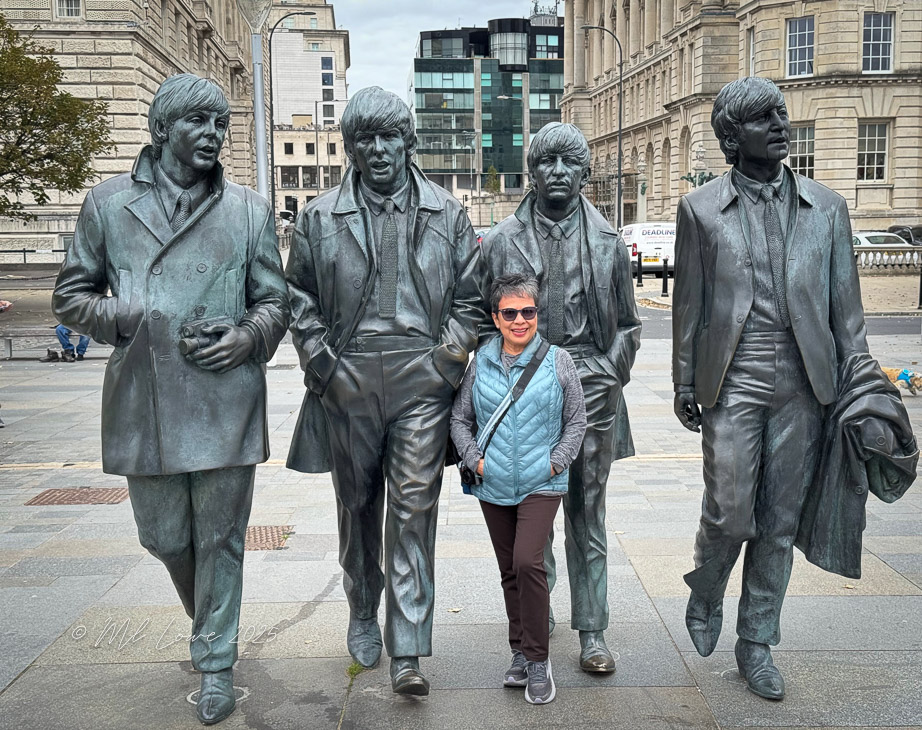 A woman stands among bronze statues of four musicians in Liverpool, England, with modern buildings in the background.