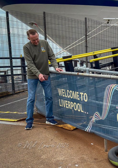 A man stands next to a banner that reads 'WELCOME TO LIVERPOOL...' at the port, with a background featuring a ship and safety fencing.