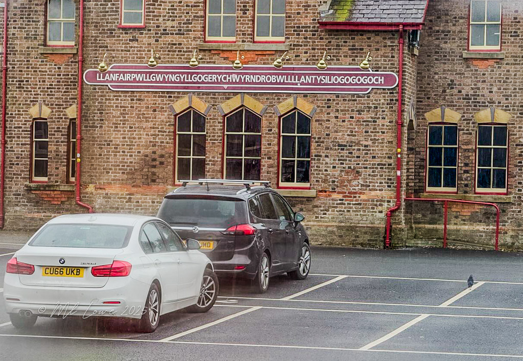 A parking lot with a white BMW and a dark vehicle in front of a brick building featuring the long Welsh name 'LLANFAIRPWLLGWYNGYLLGOGERYCHWYRNDROBWLLLLANTYSILIogogogoch' displayed prominently above.