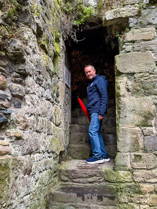 A person stands at the entrance of a stone structure with moss-covered walls, holding a red umbrella while descending a staircase.