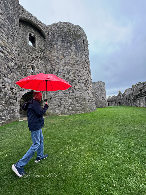 A person walking with a red umbrella near the stone walls of Beaumaris Castle on a grassy area under a cloudy sky.