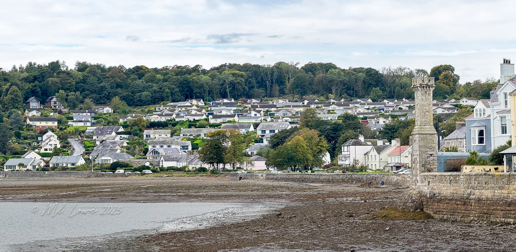 A coastal view of a quaint village in Wales, featuring a hillside of houses surrounded by greenery, with a stone tower in the foreground along the shoreline.