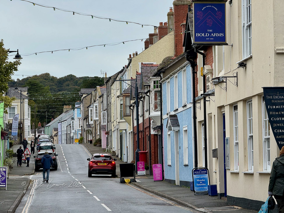 A street scene in Beaumaris, Wales, featuring colorful buildings, parked cars, and pedestrians walking along the road.