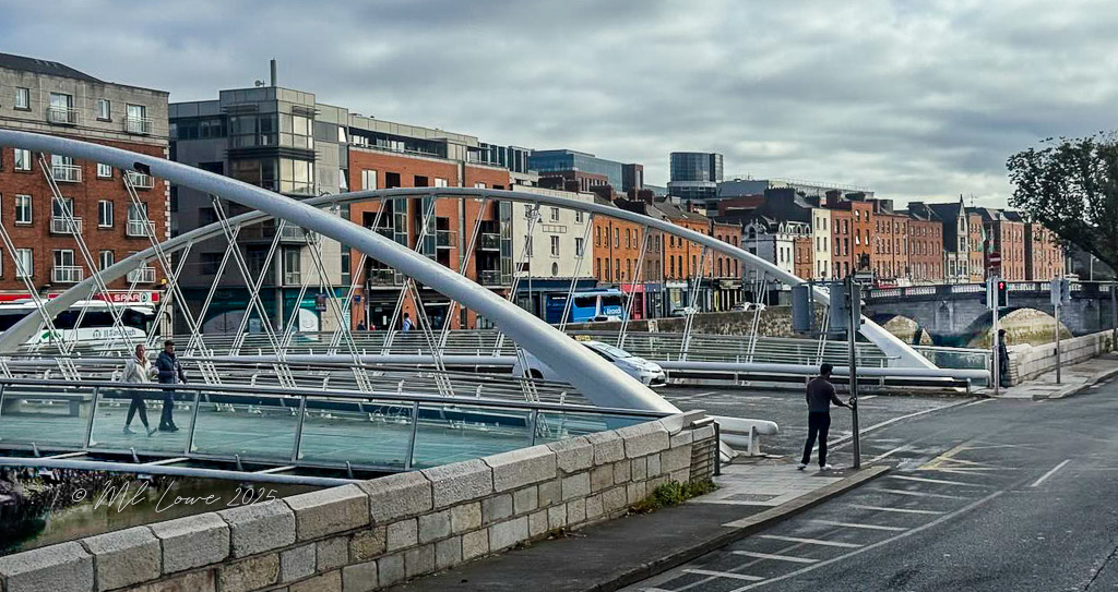 A view of the River Liffey in Dublin, featuring a modern pedestrian bridge with people walking, surrounded by colorful buildings along the riverbank under a cloudy sky.