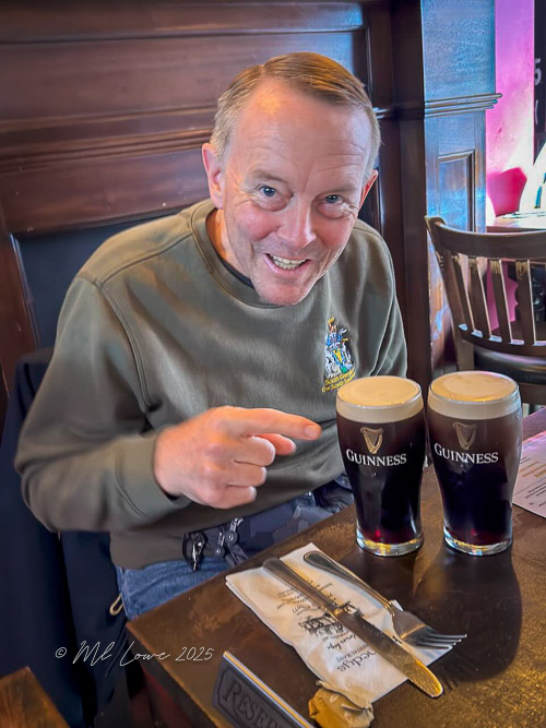 A smiling man sitting at a table in a pub, pointing at two pints of Guinness beer in front of him.
