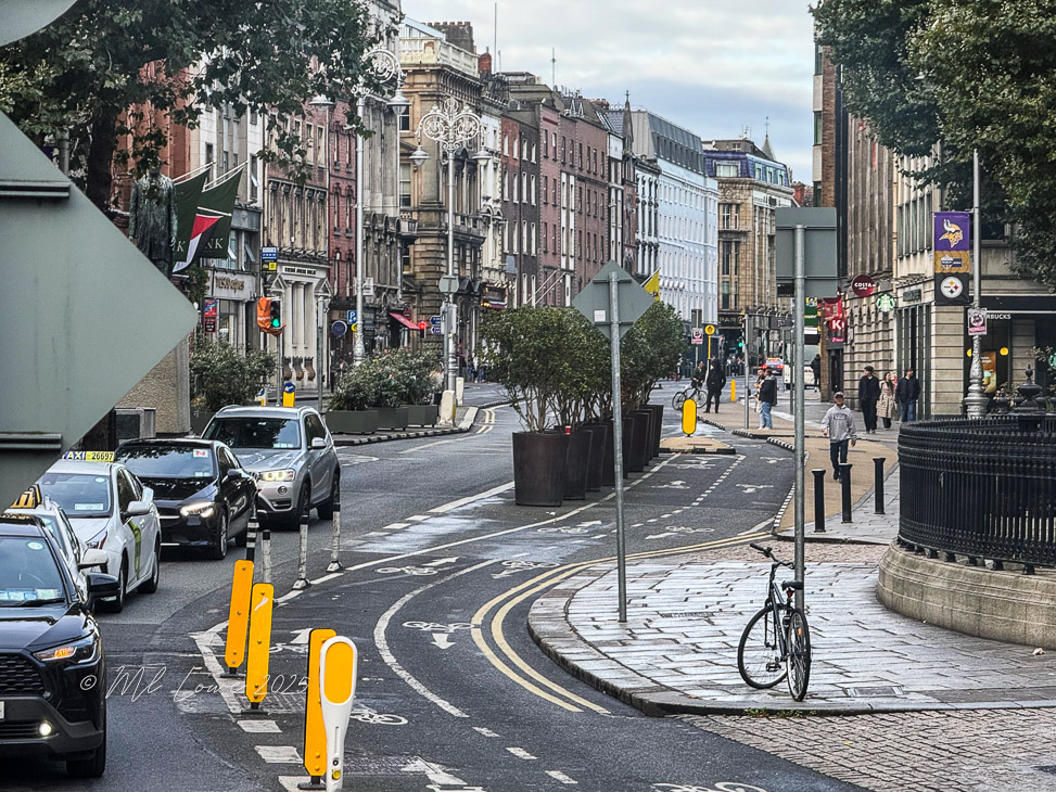 A city street in Dublin, Ireland, showcasing a mix of vehicles and pedestrians, with historic buildings lining the road and bicycle parked near a sidewalk.