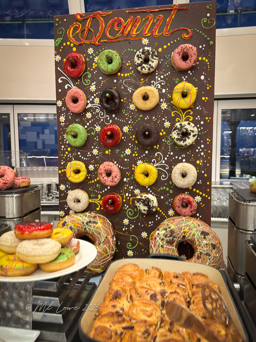 A colorful display of assorted donuts arranged on a decorative backdrop, featuring various icing colors and toppings. In the foreground, pastries are also visible.