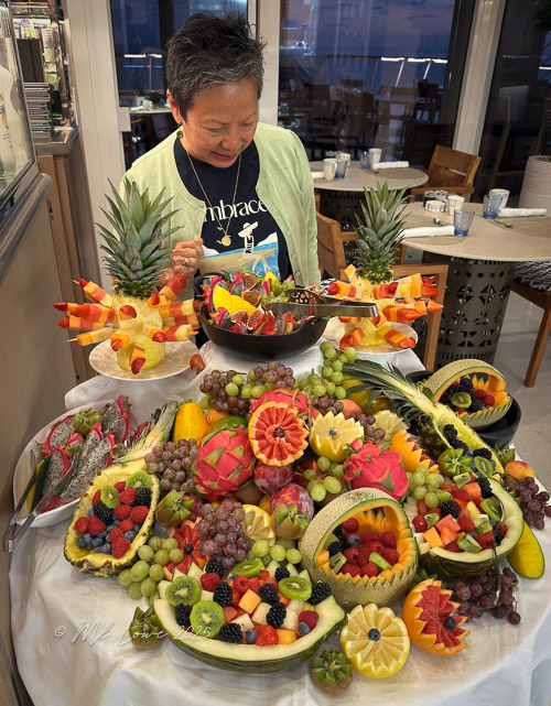 A person examines an elaborate display of creatively arranged fruit, including pineapples, melons, and various berries, showcasing a vibrant assortment of colorful and decorative fruit carvings.