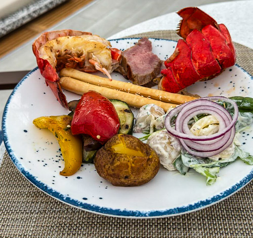 A gourmet plate featuring lobster, beef, grilled vegetables, and a side salad, served with breadsticks.