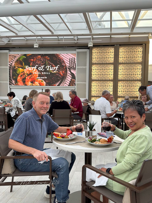 A dining couple enjoys a Surf & Turf meal at a pool grill restaurant, with a large screen displaying the menu in the background.