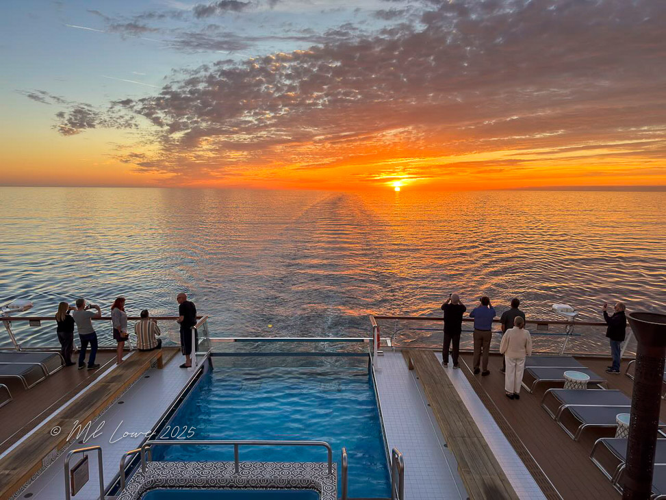 Sunset view from the deck of a cruise ship, with passengers enjoying the scenery overlooking calm waters.