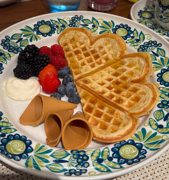 A plate featuring heart-shaped waffles drizzled with syrup, accompanied by a variety of fresh berries, a dollop of cream, and chocolate waffle cones.