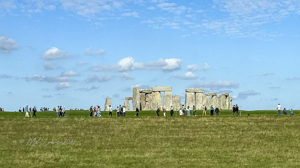 A view of Stonehenge with many visitors exploring the prehistoric monument, surrounded by grass and under a blue sky with clouds.