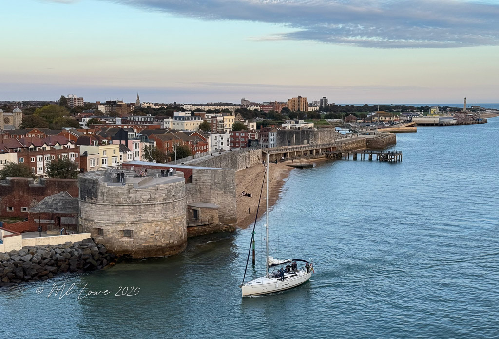 A view of a coastal town featuring a stone tower and a marina with a sailboat in the foreground, set against a backdrop of houses and a pier under a cloudy sky.