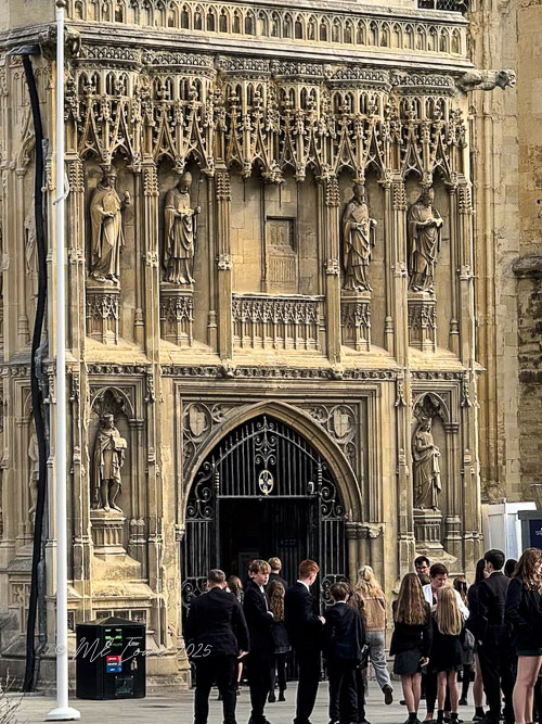Entrance of Canterbury Cathedral adorned with intricate stone carvings and statues, with a group of people dressed in formal attire gathered nearby.