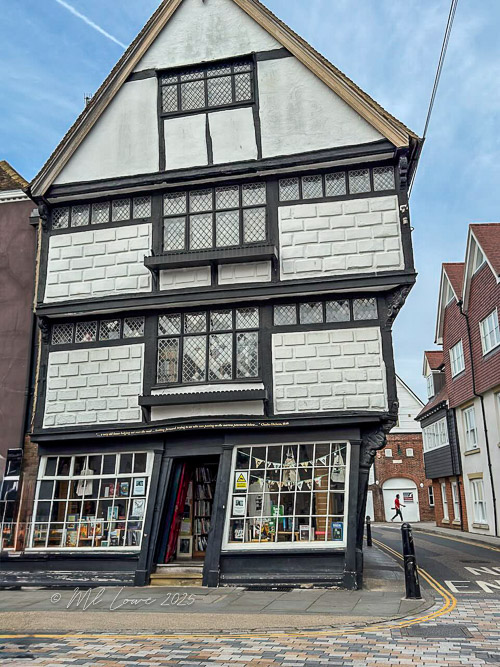 A leaning historic building with a distinct black and white design, featuring large windows and a shopfront, located on a cobblestone street.