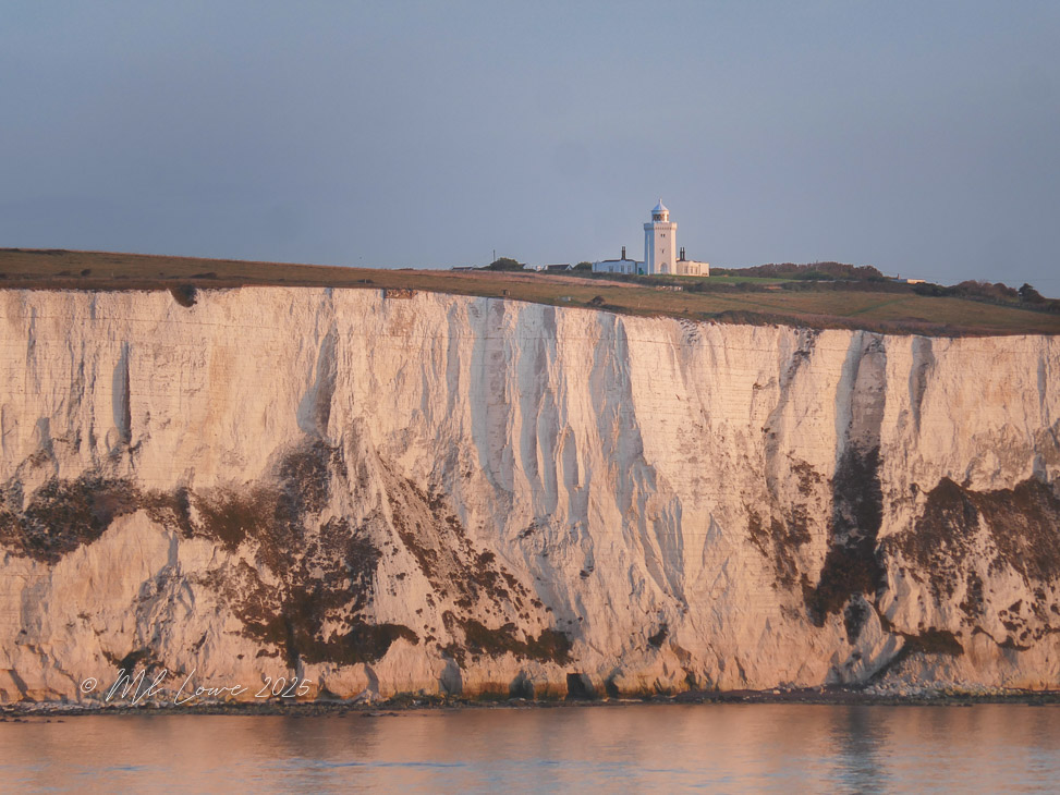 A view of the dramatic white cliffs of Dover at sunset, with a lighthouse perched on top of the cliffs overlooking the English Channel.