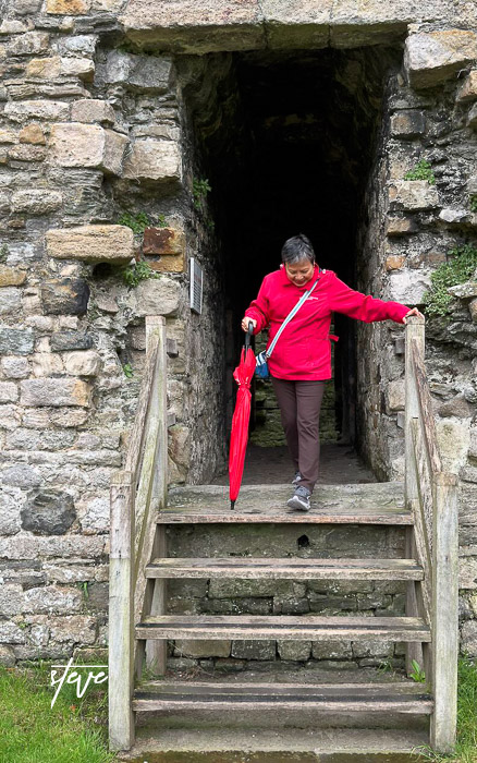 A person in a red jacket descending stone steps with an umbrella, surrounded by a historic stone structure.