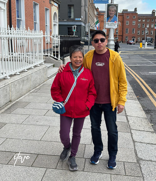 A woman in a red jacket and a man in a yellow hoodie stand together on a city sidewalk, with buildings in the background.